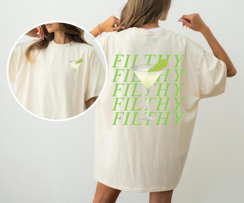 May include: Cream-colored t-shirt featuring a lime green martini glass graphic on the front and the word "FILTHY" repeated in lime green on the back. The shirt has a relaxed fit and short sleeves.