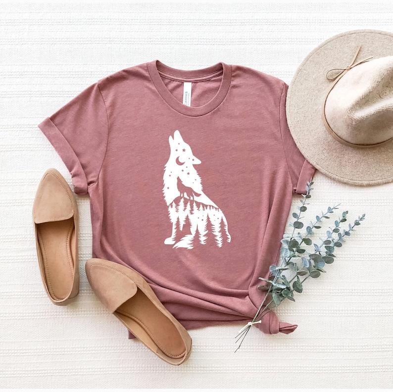 Puede incluir: Una camiseta color malva con una silueta blanca de un lobo aullando a la luna. El lobo est&aacute; rodeado de &aacute;rboles y estrellas.
