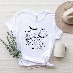 May include: White t-shirt with a black graphic of four ghosts and bats. The ghosts are in different poses and have a simple, minimalist design.