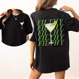 May include: Black t-shirt featuring a lime green martini glass graphic on the front and back. The back of the shirt has the word "FILTHY" repeated in a stacked design with a martini glass graphic. The shirt is a casual, oversized style.