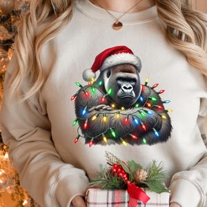May include: A cream-colored sweatshirt featuring a gorilla wearing a Santa hat and wrapped in colorful Christmas lights. The gorilla has a serious expression. A small gift box with a red bow is in the foreground.