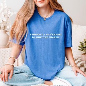 May include: A blue t-shirt with the text "I SUPPORT A MAN'S RIGHT TO SHUT THE FUCK UP" in white. The shirt is a solid color and has a crew neck. The person is wearing blue jeans.