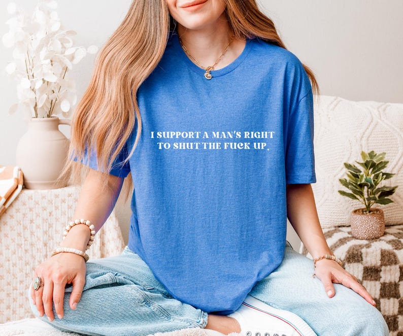 May include: A blue t-shirt with the text "I SUPPORT A MAN'S RIGHT TO SHUT THE FUCK UP" in white. The shirt is a solid color and has a crew neck. The person is wearing blue jeans.