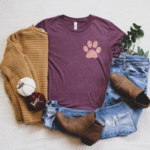 Puede incluir: Una camiseta color granate con un dise&ntilde;o de huella de pata de purpurina rosa. La camiseta est&aacute; colocada sobre una superficie blanca y peluda con un su&eacute;ter marr&oacute;n, un par de jeans azules y botines marrones.