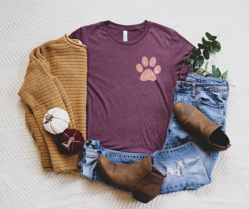 Puede incluir: Una camiseta color granate con un dise&ntilde;o de huella de pata de purpurina rosa. La camiseta est&aacute; colocada sobre una superficie blanca y peluda con un su&eacute;ter marr&oacute;n, un par de jeans azules y botines marrones.