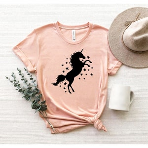 Pu&ograve; includere: Una maglietta rosa chiaro con una silhouette nera di un unicorno con un corno e una coda, circondata da stelle nere.