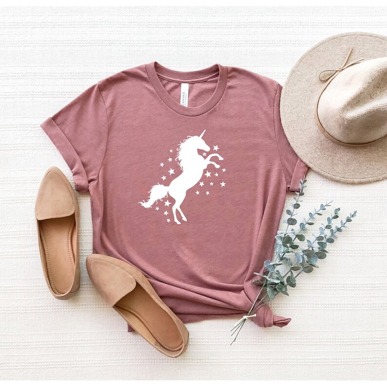 Pu&ograve; includere: Una maglietta color malva con una silhouette bianca di un unicorno con un corno e un motivo a stelle. La maglietta &egrave; stesa su una superficie bianca con un paio di ballerine marroni e un cappello di paglia.