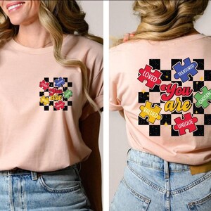 May include: A pink t-shirt with a black and white checkered background and colorful puzzle pieces that say "You are" with the words "Loved", "Valued", "Treasured", "Chosen", and "Unique" on the pieces.