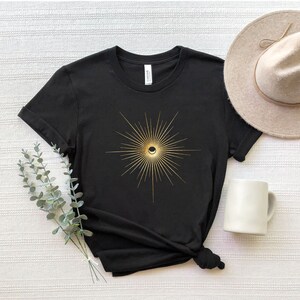 May include: Black t-shirt featuring a gold sunburst design with a crescent moon in the center. The shirt is paired with a beige hat and a white mug. The design is centered on the front of the shirt.
