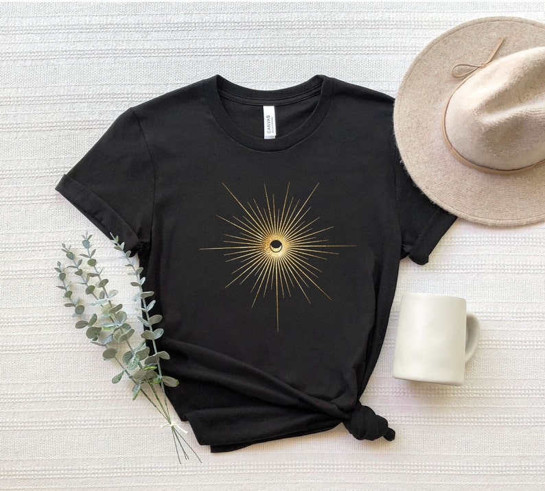 May include: Black t-shirt featuring a gold sunburst design with a crescent moon in the center. The shirt is paired with a beige hat and a white mug. The design is centered on the front of the shirt.