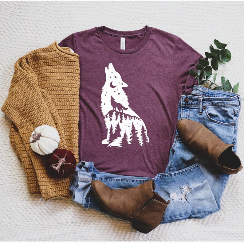 Puede incluir: Una camiseta color granate con una silueta blanca de un lobo aullando a la luna. El lobo est&aacute; rodeado de &aacute;rboles y estrellas. La camiseta est&aacute; colocada sobre una superficie blanca con un su&eacute;ter marr&oacute;n, un par de jeans azules y botas marrones.
