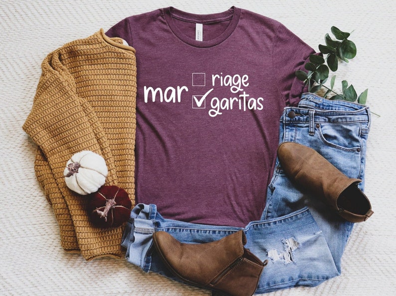 May include: A maroon t-shirt with white text that reads "mar riage margaritas" with a checkmark next to "margaritas". The shirt is styled with a brown sweater, blue jeans, and brown ankle boots.