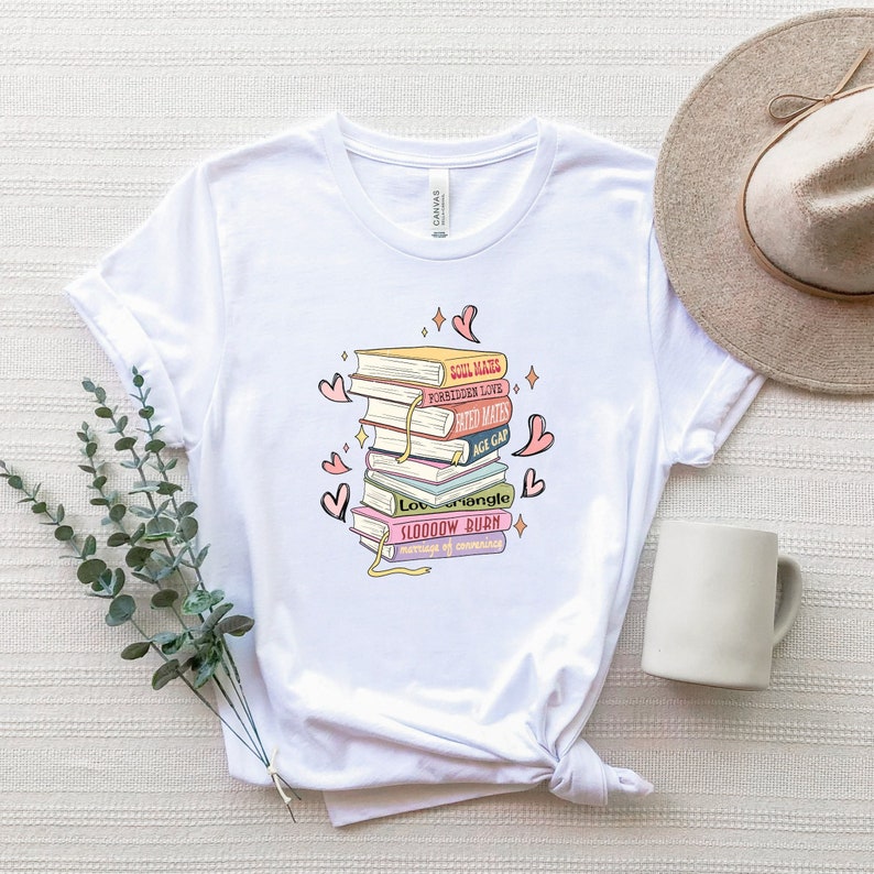 May include: White t-shirt with a graphic of stacked books, each with a title such as "Soul Mates" and "Forbidden Love." The design includes hearts and sparkles. The shirt is made of a soft material.