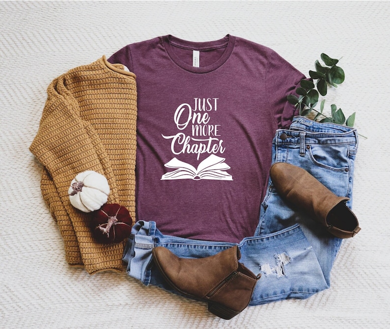 May include: A maroon t-shirt with the text "Just One More Chapter" in white, featuring a stylized open book graphic. The shirt is displayed on a white surface with a brown sweater, blue jeans, and brown boots.