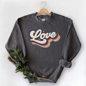 Puede incluir: Una sudadera gris oscuro con un gr&aacute;fico de "Love" de estilo retro en azul, naranja, amarillo y blanco.