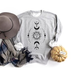 Puede incluir: Una sudadera gris con un dise&ntilde;o negro de sol y luna. El sol tiene una cara y est&aacute; rodeado por un c&iacute;rculo de lunas.
