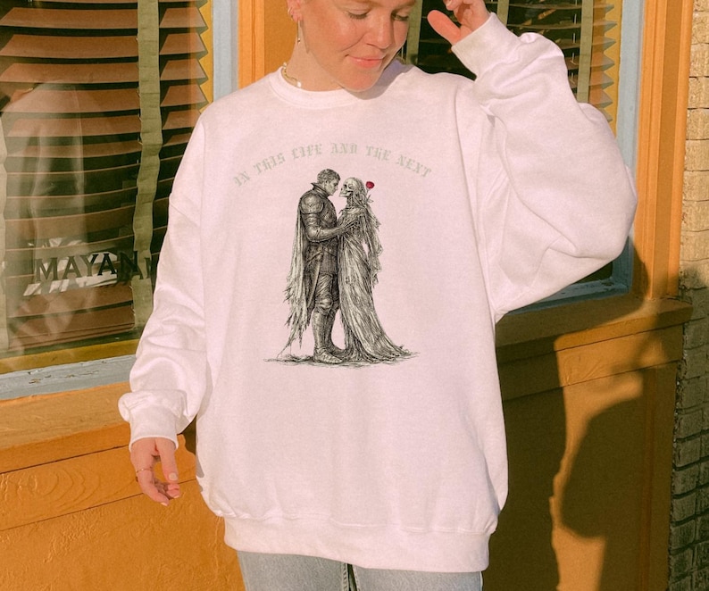 May include: White sweatshirt featuring a black and white illustration of a skeletal couple embracing. The text "IN THIS LIFE AND THE NEXT" is printed above the illustration. The sweatshirt is a casual, comfortable style.