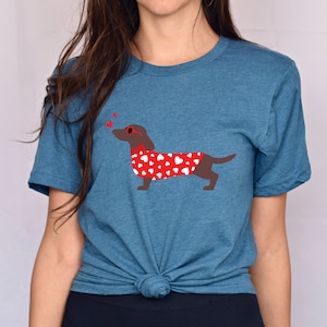 Puede incluir: Una camiseta azul turquesa con un dachshund marr&oacute;n que lleva un su&eacute;ter rojo con corazones blancos y gafas de sol en forma de coraz&oacute;n rojo. Tres corazones rojos flotan sobre el dachshund.