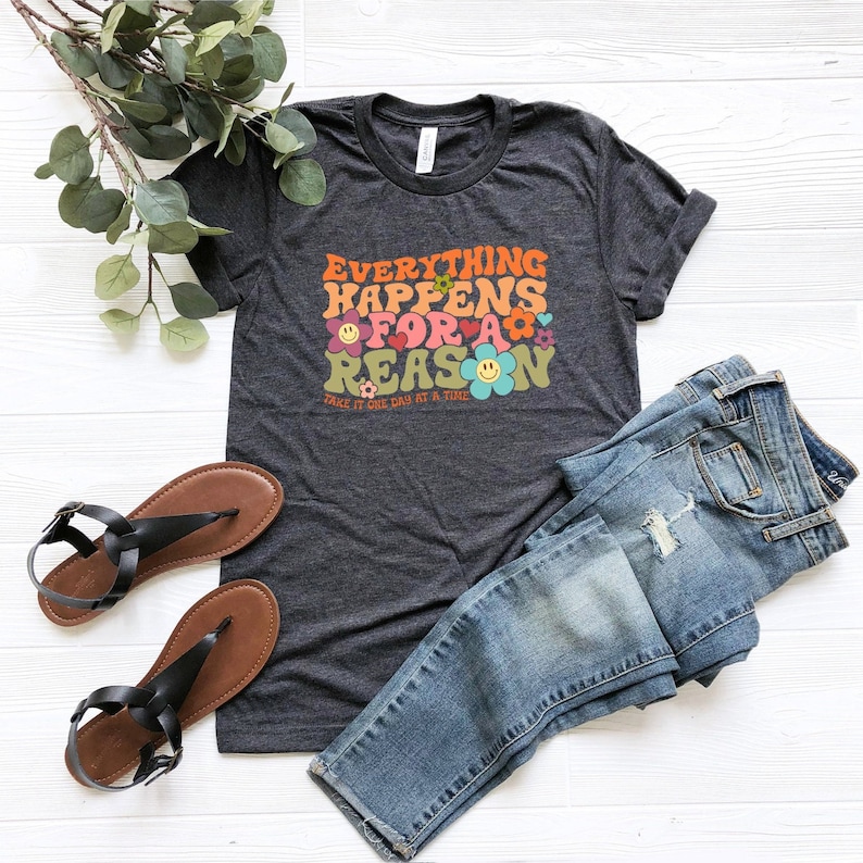 Puede incluir: Una camiseta gris oscuro con el texto "Everything Happens For A Reason" en letras coloridas de estilo retro. El texto est&aacute; rodeado de flores y caras sonrientes. El texto "Take it one day at a time" est&aacute; impreso debajo del texto principal.