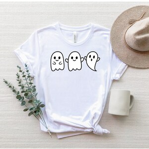 May include: White t-shirt with a graphic of three cute ghosts. The ghosts are in a row and have different expressions. The shirt is a crew neck style and has a relaxed fit.