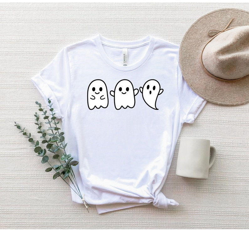 May include: White t-shirt with a graphic of three cute ghosts. The ghosts are in a row and have different expressions. The shirt is a crew neck style and has a relaxed fit.