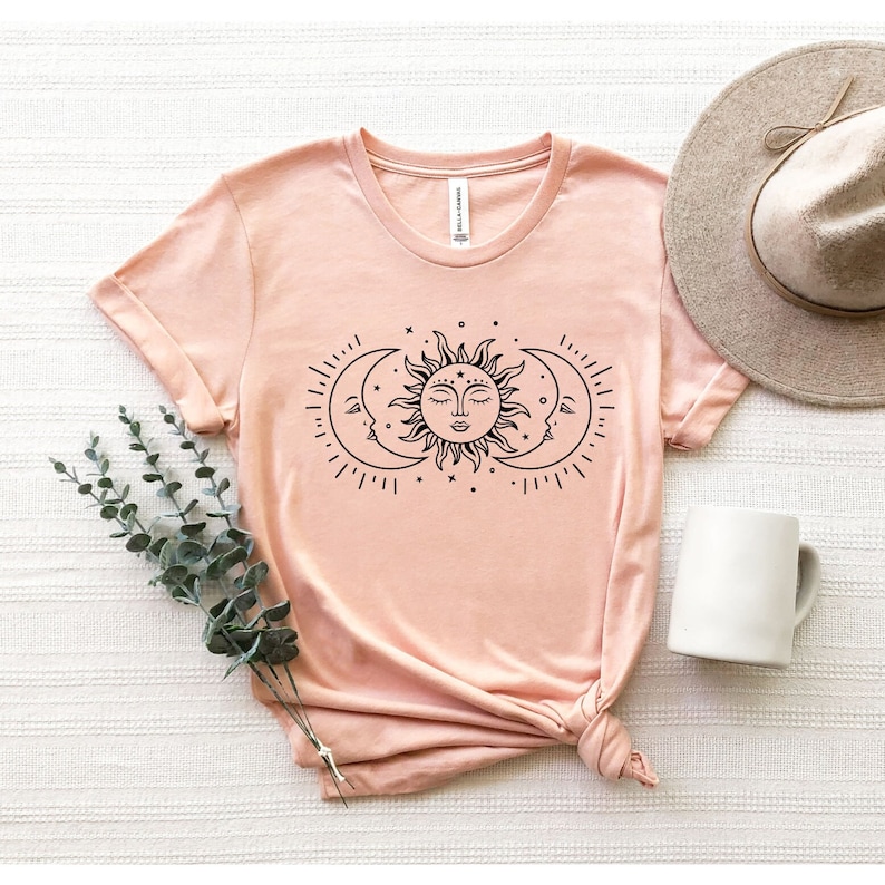 May include: A peach-colored t-shirt with a black graphic of a sun and two moons. The sun has a face and the moons are crescent shaped.