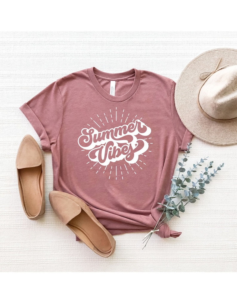 May include: A mauve colored t-shirt with the text "Summer Vibes" in a white retro font. The shirt is laid flat on a white surface with two brown loafers and a straw hat.