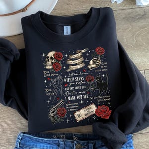 May include: Black sweatshirt with a graphic design featuring a skull, roses, a hand, a cat, and a gun. The design includes text that reads "Let me know which stars you prefer" and "House of Mirrors."