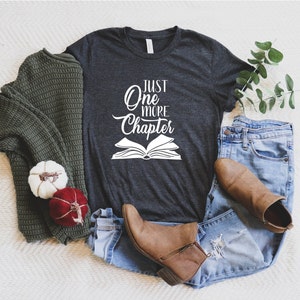 May include: A dark gray t-shirt with the text "Just One More Chapter" in white lettering and a white outline of an open book.