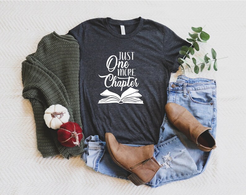 May include: A dark gray t-shirt with the text "Just One More Chapter" in white lettering and a white outline of an open book.