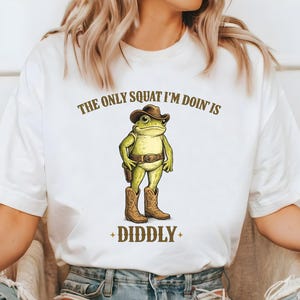May include: White t-shirt featuring a cartoon frog dressed as a cowboy, complete with a hat, boots, and a holster. The text above reads "THE ONLY SQUAT I'M DOIN' IS" and below "+ DIDDLY +" in brown lettering.