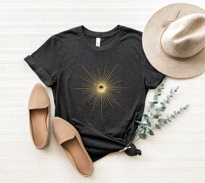 May include: Dark gray t-shirt with a gold sunburst design featuring a crescent moon. The shirt is styled with beige flats and a wide-brimmed hat. A sprig of greenery adds a natural touch to the display. The shirt is made of a soft material.