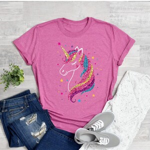 以下が含まれることがあります： キラキラ光るユニコーンのグラフィックがプリントされたピンクのTシャツ。ユニコーンはピンク、ブルー、イエロー、パープルで、金色の角をしています。Tシャツはクルーネックで、袖は短いです。