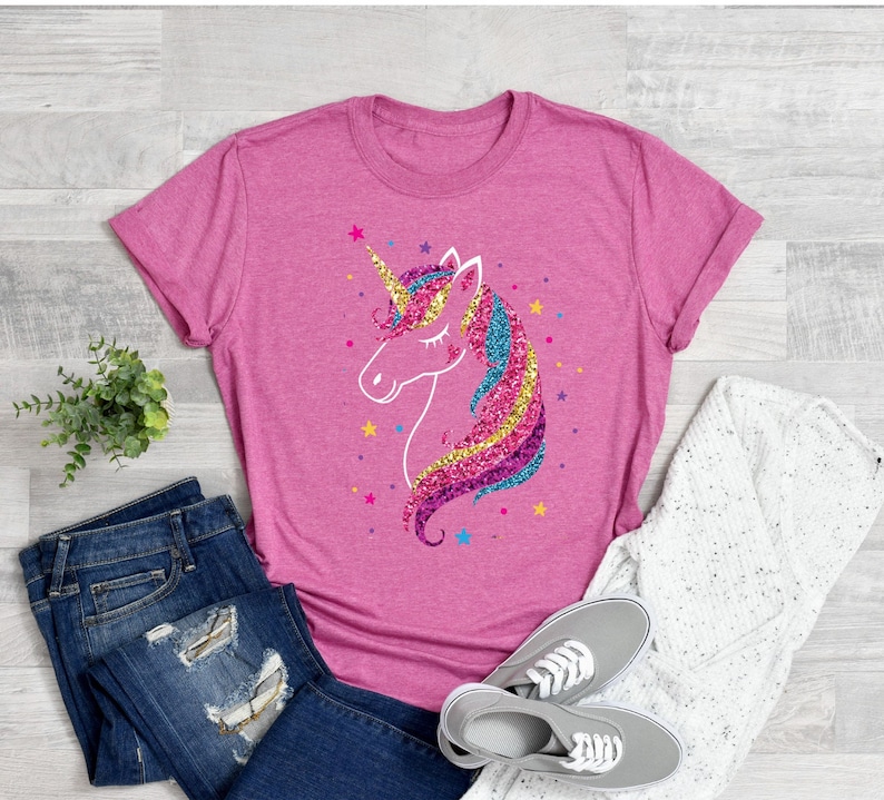 以下が含まれることがあります： キラキラ光るユニコーンのグラフィックがプリントされたピンクのTシャツ。ユニコーンはピンク、ブルー、イエロー、パープルで、金色の角をしています。Tシャツはクルーネックで、袖は短いです。