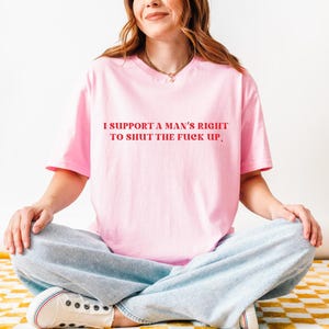 May include: A light pink t-shirt with the text "I SUPPORT A MAN'S RIGHT TO SHUT THE FUCK UP" in red. The shirt is worn by a person sitting cross-legged, wearing light blue jeans and white sneakers.