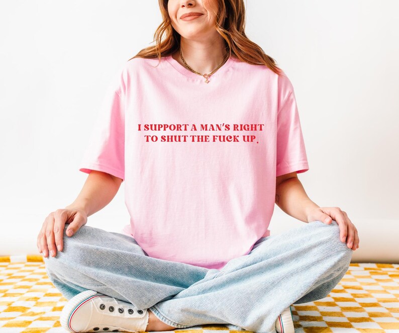 May include: A light pink t-shirt with the text "I SUPPORT A MAN'S RIGHT TO SHUT THE FUCK UP" in red. The shirt is worn by a person sitting cross-legged, wearing light blue jeans and white sneakers.