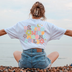May include: A white t-shirt with a colorful graphic that says "Stay Wild Moon Child" in a retro font. The shirt is being worn by a person sitting on a beach.