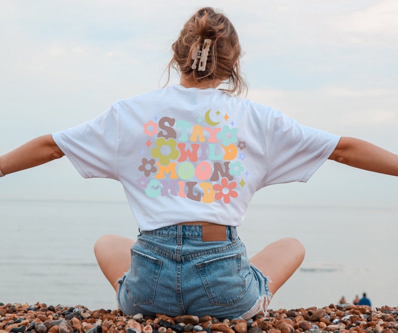 May include: A white t-shirt with a colorful graphic that says "Stay Wild Moon Child" in a retro font. The shirt is being worn by a person sitting on a beach.