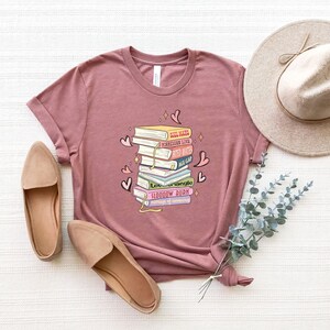 May include: Mauve t-shirt with a graphic of stacked books, including titles such as "Soul Mates" and "Forbidden Love." The design incorporates hearts and stars. The image also shows tan flats and a beige hat.