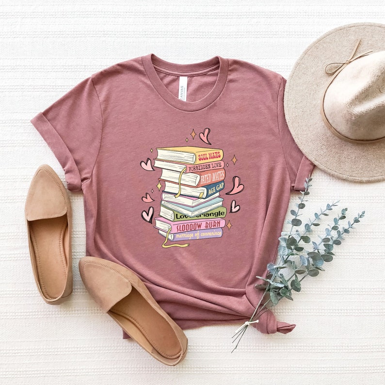 May include: Mauve t-shirt with a graphic of stacked books, including titles such as "Soul Mates" and "Forbidden Love." The design incorporates hearts and stars. The image also shows tan flats and a beige hat.