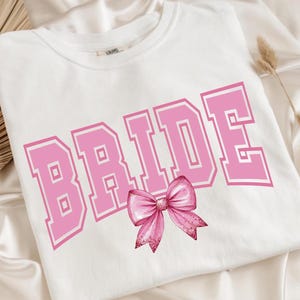 May include: White t-shirt with the word "BRIDE" printed in pink with a pink bow underneath.