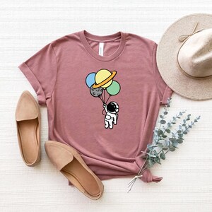 May include: A mauve t-shirt with a graphic of an astronaut holding onto a bunch of planets as balloons. The planets are yellow, blue, green, and gray.