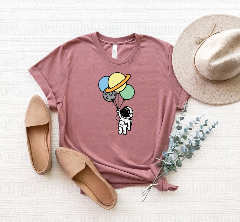 May include: A mauve t-shirt with a graphic of an astronaut holding onto a bunch of planets as balloons. The planets are yellow, blue, green, and gray.