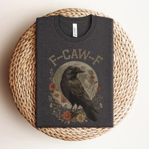 May include: Dark gray t-shirt featuring a black raven illustration with the text "F-CAW-F". The raven is set against a full moon and surrounded by flowers in shades of red, yellow, and green. The shirt is displayed on a woven surface.