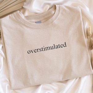May include: A beige t-shirt with the word "overstimulated" printed in black. The shirt is folded on a white satin surface, with dried plants and a fan-shaped object in the background.