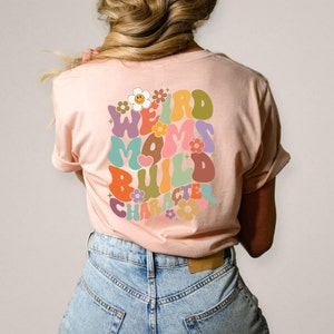 May include: A pink t-shirt with a retro-style graphic that says "Weird Moms Build Character" in colorful, rounded letters with flowers surrounding the text.