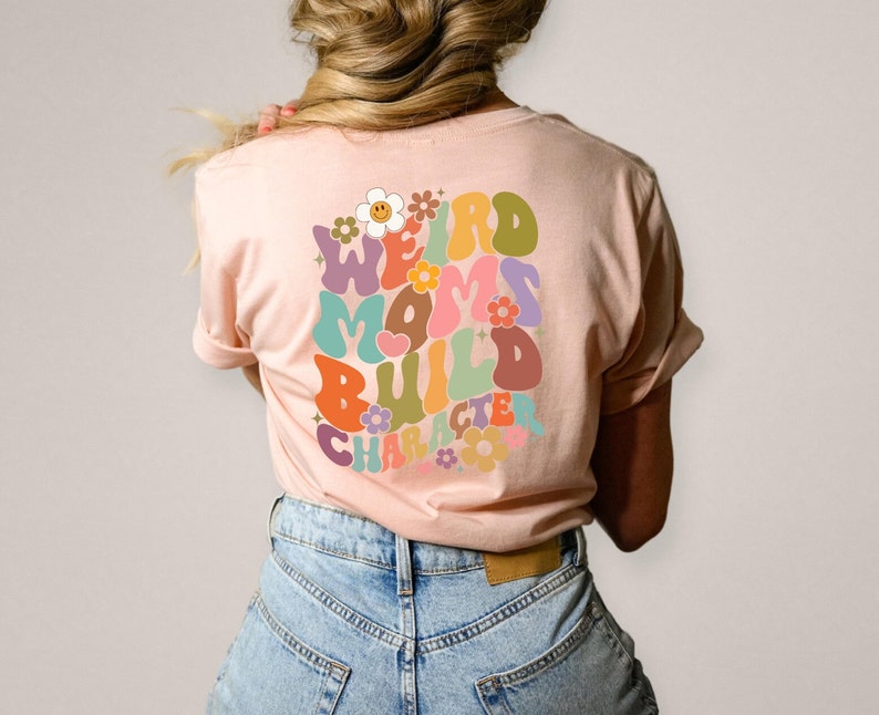 May include: A pink t-shirt with a retro-style graphic that says "Weird Moms Build Character" in colorful, rounded letters with flowers surrounding the text.
