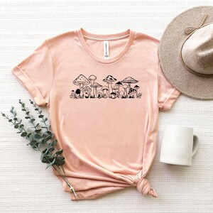 以下が含まれることがあります： 薄いピンクのTシャツに、黒いキノコの列のグラフィックがプリントされています。このTシャツは、柔らかく快適な素材で作られています。