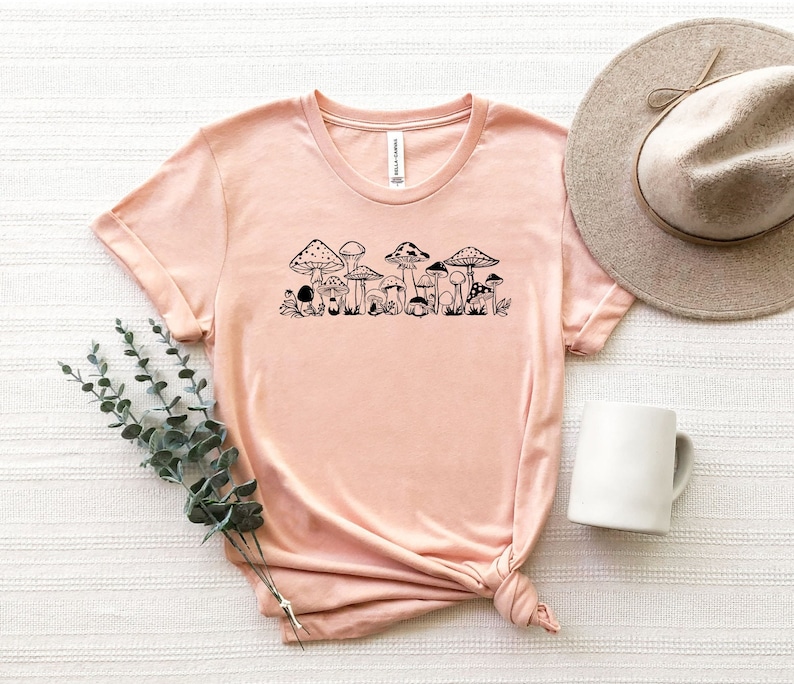 以下が含まれることがあります： 薄いピンクのTシャツに、黒いキノコの列のグラフィックがプリントされています。このTシャツは、柔らかく快適な素材で作られています。