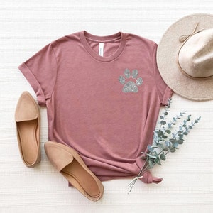 Puede incluir: Una camiseta color malva con un dise&ntilde;o de huella de pata de purpurina plateada. La camiseta est&aacute; colocada sobre una superficie blanca con un par de zapatos planos beige y un sombrero de paja.